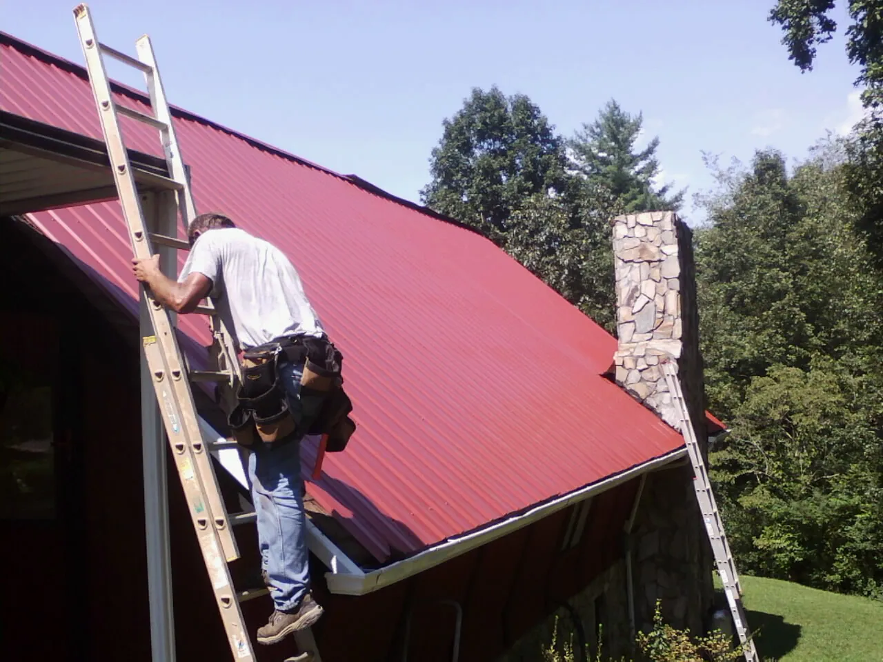 Roofer on a ladder beside a red metal roof and stone chimney — Soffit & Fascia Repair by Roofing Brothers in Washington Crossing